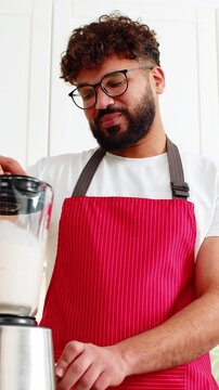 Arabian man blending whey protein smoothie with banana apple after workout in home kitchen. Young guy smiling using blender for healthy sports nutrition drink for muscle recovery in small apartment