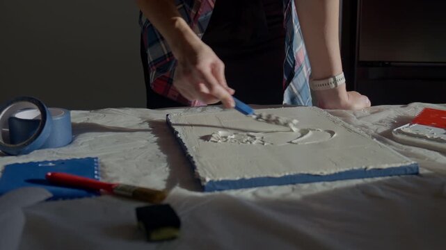 Woman applying textured paste to canvas using palette knife on work table with art tools. Concept of creative process, handmade artwork