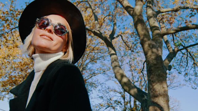 Attractive Caucasian blonde woman in black hat, sunglasses and coat. Portrait of a positive stylish lady against tree in autumn.
