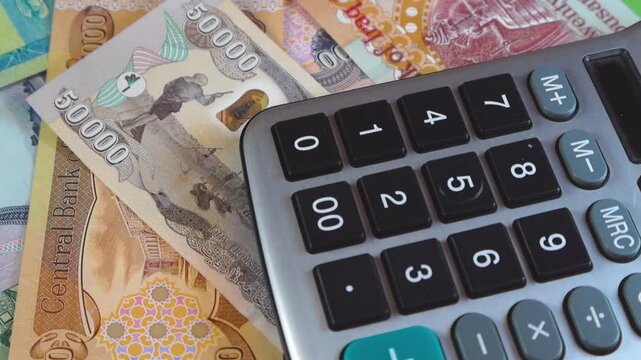 A camera pans over a scattered pile of Iraqi Dinar banknotes (50k, 25k, 10k, 5k) alongside a black calculator. The shot emphasizes financial calculation, currency exchange, and Iraqi heritage.