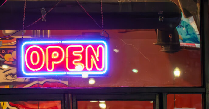 Vibrant pink and blue neon 'OPEN' sign glowing brightly in a storefront window at night, reflecting urban light and welcoming customers into a business.