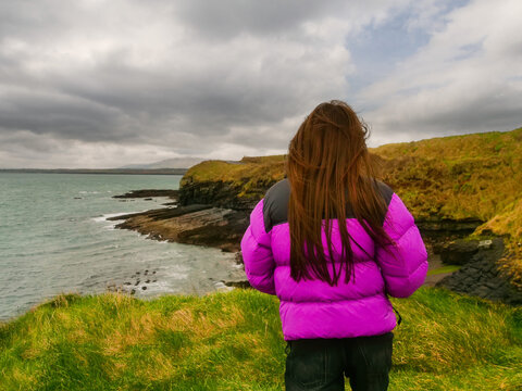 A woman stands on a grassy hill overlooking the ocean and cliffs. She is wearing a purple jacket and black pants. Mullaghmore area in county Sligo, Ireland. Travel and tourism. Cloudy sky.