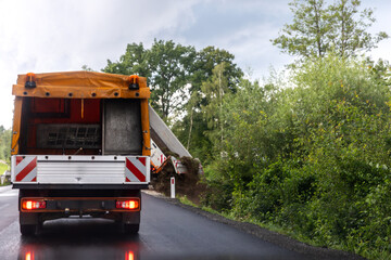 Roadwork truck warning lights drives along wet asphalt road near green vegetation countryside landscape rain. Maintenance vehicle operates roadside clearing soil debris after storm weather conditions © Kyryl Gorlov