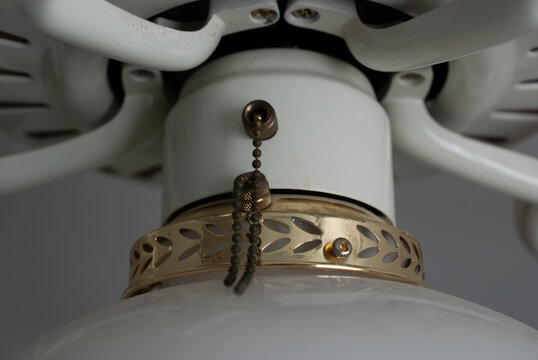 Close-up of a vintage-style white ceiling fan light fixture with decorative brass accents and functional pull chains, highlighting minor dust buildup and classic home interior design details.