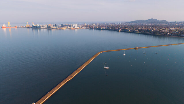 Panoramic view of the Edinburgh coastline looking from Granton towards the Port of Leith. The scene features a long historical stone pier stretching across the blue water of the Firth of Forth. Backgr
