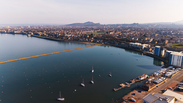 Sea view of Edinburgh skyline and Granton harbor with sailing boats and yachts. Scenic maritime panorama featuring traditional stone architecture, a long pier and residential buildings along the Firth