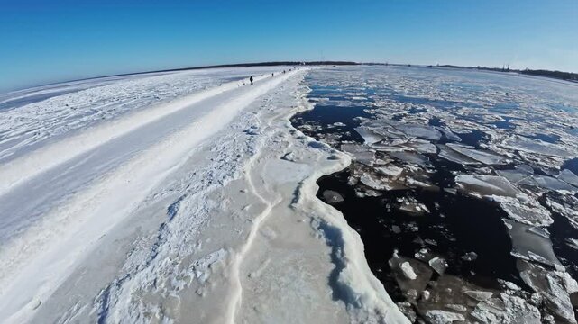 Aerial follows a snow covered breakwater toward a small lighthouse on the Riga coastline. Ice ridges and open water appear as walkers move under crisp midday light.