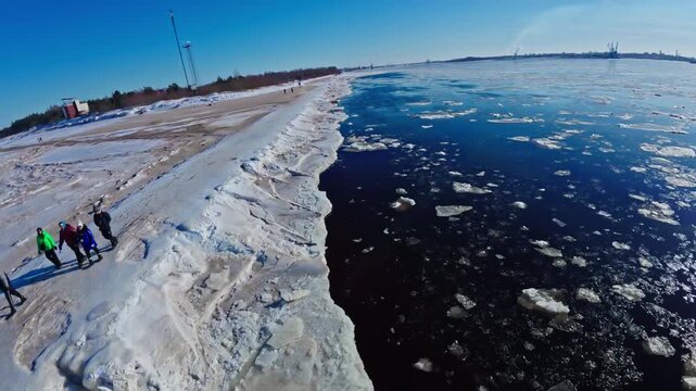 Aerial drone track follows jagged ice and icy berm by sandy snowy beach near the Daugava River mouth in Riga, with small lighthouse and distant port cranes visible.