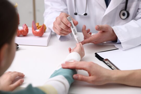 Endocrinologist checking little girl's blood sugar level with lancet pen while her mother supporting her at white desk indoors, closeup