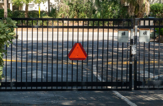 Automatic black metal security gate with a bright orange slow-moving vehicle sign and prominent warning signs, providing controlled access to a residential community entrance on a sunny day.