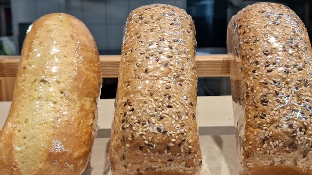 Loaves of freshly baked bread wrapped in transparent plastic film on wooden bakery shelf. Variety of artisanal bread including sesame and flax seeds. Wrap protects fresh crust golden texture