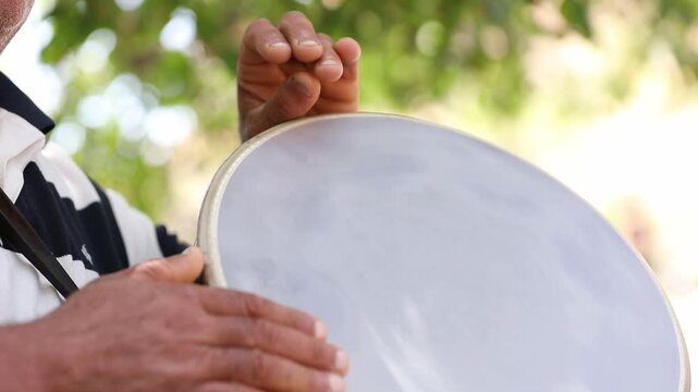 Close up shot of an older man's hands rhythmically playing a traditional darbuka hand drum outdoors against a blurred green background of trees on a bright sunny day.