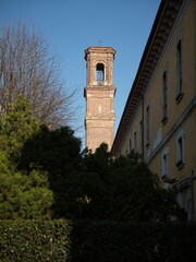 Obraz premium View of a historic Italian bell tower rising above trees beside a classical building. Warm sunlight enhances textures and architectural details against a clear blue sky.