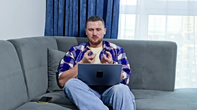 A person is seated on a grey sofa in a living room. He is looking at a laptop screen while holding two small blue items in his hands. A mobile phone rests nearby.
