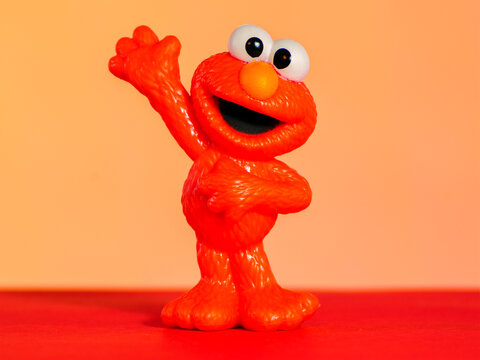 Utica, NY - Jan 6, 2026: An Elmo figurine from Sesame Street, shown standing with one arm raised and the other on its belly, photographed on a red surface with an orange gradient background