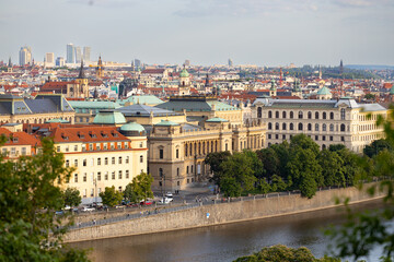 Fototapeta premium Aerial view of historic buildings along the Vltava River in Prague, showcasing architectural styles and greenery with city skyline in the background