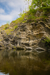 Fototapeta premium Rocky cliffside with lush greenery reflecting in calm water under a partly cloudy sky in a natural outdoor setting