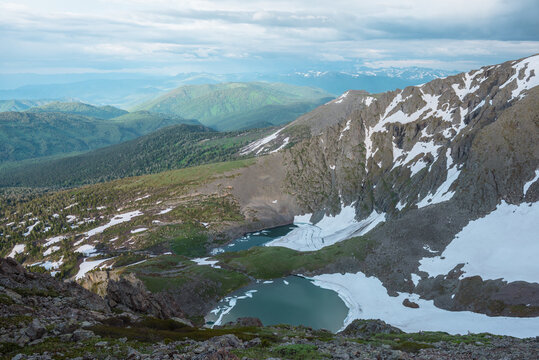 Awesome harsh top view from rock precipice edge to two turquoise alpine lakes with ice floes in stone mountain cirque. Small creek between two green icy lakes in rocky stony terrain under cloudy sky.