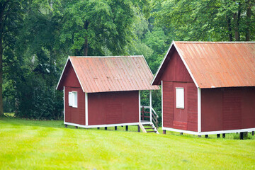 Fototapeta premium Two red wooden small houses with sloped roofs situated on a green grassy field surrounded by trees in a serene rural landscape