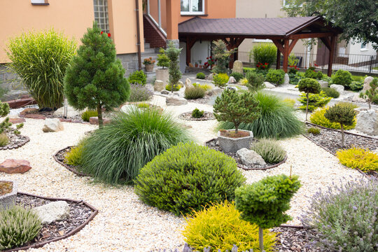 Japanese-style garden featuring various shrubs, ornamental stones, and gravel pathways, with a wooden pavilion and colorful plants in the background