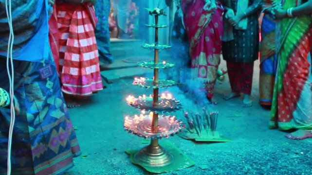 Close-up of a traditional multi-tier brass oil lamp (Deepam or Diya) illuminated during a Hindu religious ceremony in India. Surrounded by devotees in colorful sarees