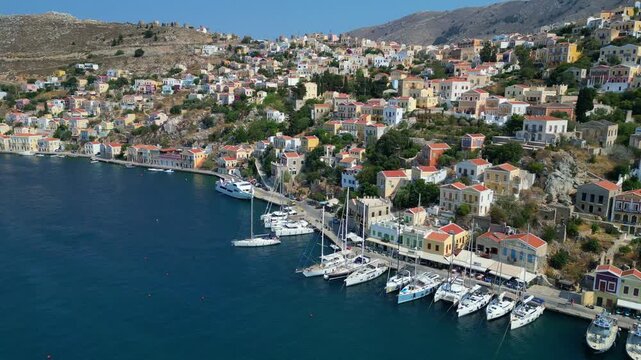 Aerial panorama of colorful neoclassical houses and luxury yachts in Symi harbor, Greece. Picturesque Dodecanese island coastline at sunny day, 4K drone footage