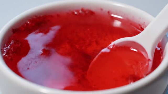 Red powder dissolving in clear water being stirred with a white spoon inside a white ceramic bowl close up