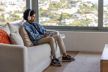 Woman sitting on sofa by window holding tablet, wearing denim jacket, headphones, copy space