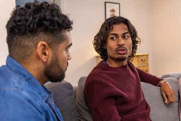 Indian men sitting on gray sofa in living room talking while wearing maroon sweater, denim shirt