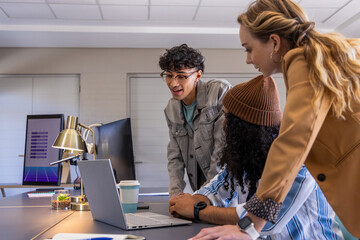 Coworkers collaborating around laptop in meeting room, denim jacket and beanie viewing purple graph