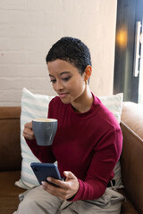 African American woman sitting on leather couch wearing burgundy sweater holding phone and gray mug