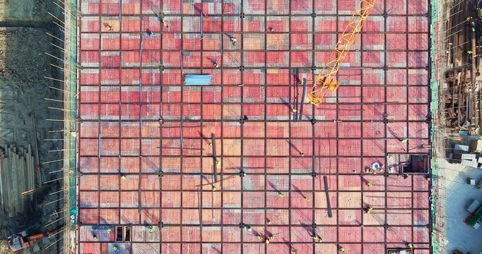 Top down aerial view of construction workers installing steel rebar grid and formwork for large building foundation slab at urban development site.