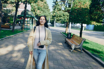 Fototapeta premium Woman walking along a park path with a camera hanging from her neck, wearing a light trench and jeans, enjoying outdoor leisure among trees and benches