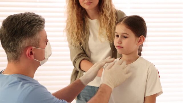 Endocrinologist in medical gloves and protective mask examining girl's thyroid gland while her mother supporting her in clinic. Camera moving forward