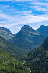 Obraz premium Dense evergreen forest fills a mountain valley with a winding river leading toward rugged peaks under a blue sky in Glacier National Park, Montana, USA.
