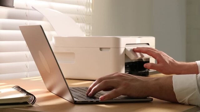 Man using printer while working on laptop at wooden desk indoors, closeup