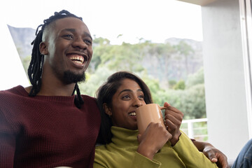 Diverse couple sitting closely in knit sweaters on sofa by sliding glass door, holding beige mug