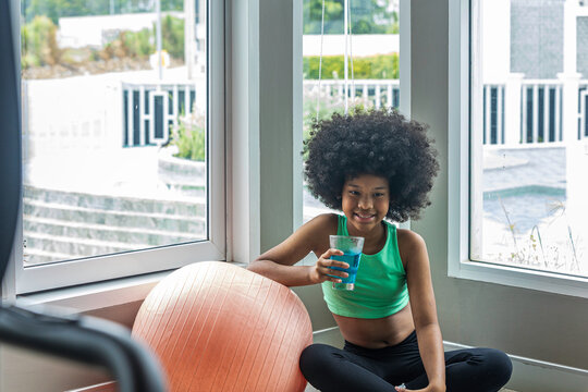 African teen girl with natural hair relaxing after a workout, sitting by a large window and drinking a replenishing electrolytes, representing healthy recovery, active lifestyle, and youthful energy