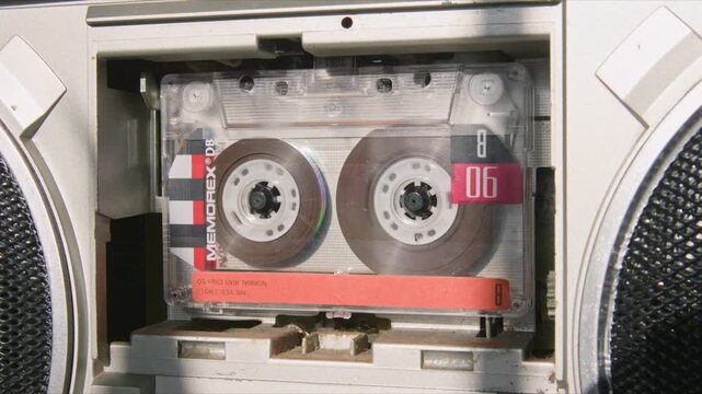 Macro footage: vintage 1980s cassette spins in boombox player. Tape reels turn, old hits from the 80s play, warm lighting highlights classic analog music device.