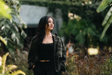 Young woman black hair casual jacket outdoor garden greenery natural light smiling relaxed confident standing foliage nature lifestyle portrait happiness beauty fashionable trendy serene peaceful © chokniti