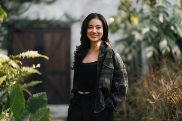 Naklejka premium Young woman black hair casual jacket outdoor garden greenery natural light smiling relaxed confident standing foliage nature lifestyle portrait happiness beauty fashionable trendy serene peaceful