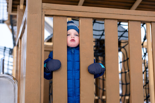 A child in warm winter clothing stands on a snowy playground beside a rope tunnel