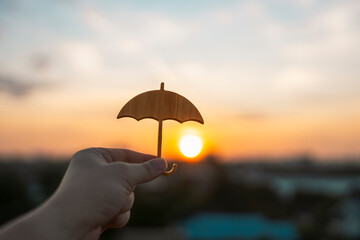Hand holding small wooden umbrella icon against sunset sky. Concept of protection, security, insurance coverage, financial safety, and life guarantee with warm evening light. © sommart