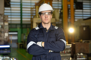 Confident man factory worker in an industrial portrait stands serious with arms crossed wearing safety helmet uniform and glasses feeling professional and proud inside manufacturing plant © 220 Selfmade studio