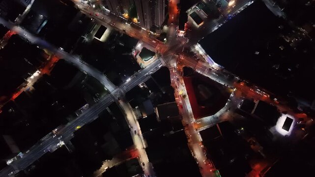 Night top-down descending drone captures a complex multi-road junction where five or six roads converge at sharp angles creating an abstract star pattern of amber-lit arteries between dark blocks