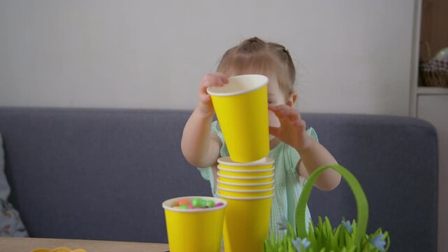 Little girl stacks yellow cups with care. She smiles as colorful candies rest inside. Soft light highlights her playful, focused expression. Perfect for Easter, child development, or family moments