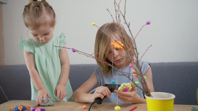 Two young girls craft colorful decorations on branches. They focus intently, using glue and small beads. Tools and supplies lie nearby on a wooden table. Children enjoy creative