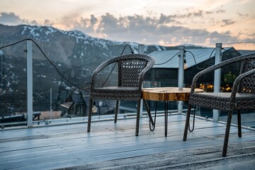 Fototapeta premium A serene outdoor seating area featuring two wicker chairs and a wooden table, set against a backdrop of mountains and a beautiful sunset sky.