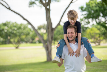 Fototapeta premium Smiling father carrying young son on his shoulders while playing together outdoors in public park. Joyful man giving boy shoulder ride in nature family bonding moment outdoor fun on summer.
