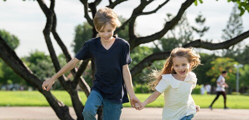 Two happy children boy and girl with smile holding hands and running together in public park....
